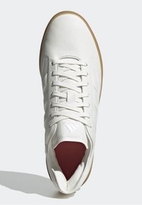 Vit sneaker med en texturerad läderöverdel, platta snören och en brun gummisula. Har en logotyp på tungan och en färgad insida.