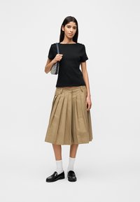 Μαύρο τοπ με κοντά μανίκια, μπεζ πλισέ μίντι φούστα, λευκές αθλητικές κάλτσες και μαύρα μοκασίνια. Το μοντέλο κρατάει μια τσάντα με σχέδια, με ουδέτερο φόντο.