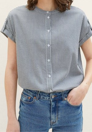 Chemise à rayures bleu marine et blanche, à manches courtes, sans col, en tissu doux avec une coupe déstructurée, portée avec un jean bleu.