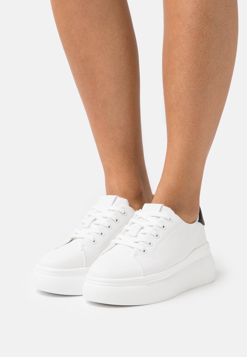 Zapatillas de cuero blancas con puntera redondeada, cordones planos y suela de goma elevada. Acento negro en el talón; textura suave en general.