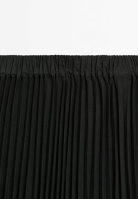 Détail d'une jupe plissée noire avec une ceinture élastique froncée et de fines plis verticaux sur un fond clair uni.