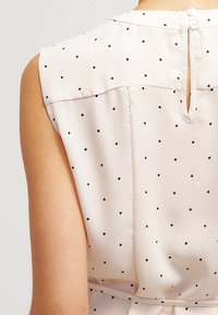 Blusa senza maniche bianca con pois neri, chiusura sul retro con bottoni e una piccola apertura a forma di chiave nella parte superiore della schiena.