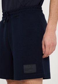 Mørkeblå bomuldsbukser med elastisk talje, prydet med et sort gummilogo-patch og glat tekstur. Enkel design uden lommer.