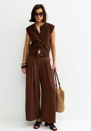 Vrouw met een bruin mouwloos hemd met knopen en wijde broekspijpen, draagt een gevlochten boodschappentas, een zonnebril en zwarte sandalen.