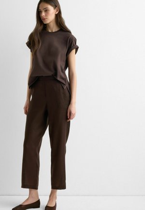 Frau trägt ein dunkelbraunes kurzärmliges Oberteil und passende weit geschnittene Hosen, steht vor einer schlichten weißen Wand.