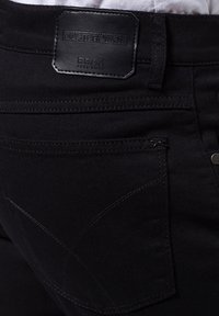 Černé džínové kalhoty s koženou nášivkou, zdobené složitými švy a kapsou na zip, které prezentují elegantní, moderní design.