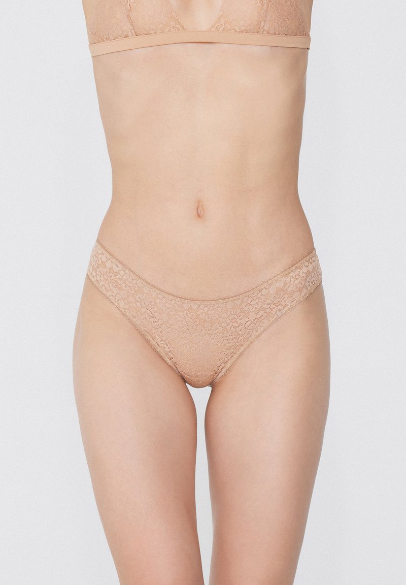 Femme portant un soutien-gorge en dentelle nude et une culotte en dentelle assortie, vue du milieu du torse jusqu'à la partie supérieure des cuisses sur fond blanc.
