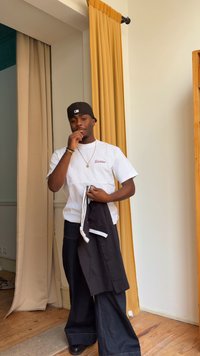 Joven con gorra negra, camiseta blanca Dickies y vaqueros anchos sosteniendo una chaqueta negra, de pie junto a cortinas beige y amarillas en el interior.