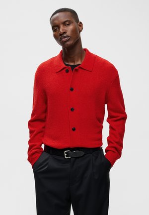 Homme les mains dans les poches, portant un pull rouge boutonné et un pantalon noir avec ceinture, sur fond clair uni.