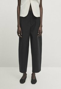 Czarne spodnie szerokie wykonane z lekkiego materiału, o prostym kroju, wysokim stanie i delikatnym drapowaniu. W połączeniu z plecionymi czarnymi butami.