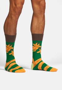 Grüne Socken mit einem Giraffenmuster, versehen mit orangen Akzenten, braunen gerippten Bündchen und unterschiedlichen Streifen in Orange und Grün.