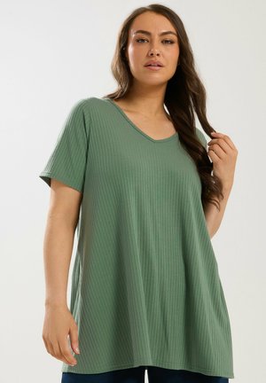 Frau mit langen braunen Haaren, trägt ein locker sitzendes, geripptes Kurzarmoberteil in Salbeigrün, berührt ihr Haar, neutraler Hintergrund.