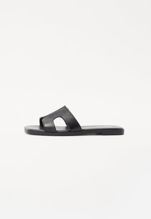 Sort lædersandal med slideryg, der har et udsagnsfuldt skæremønster, flad sål og glat overflade. Simpelt design uden dekorative detaljer.
