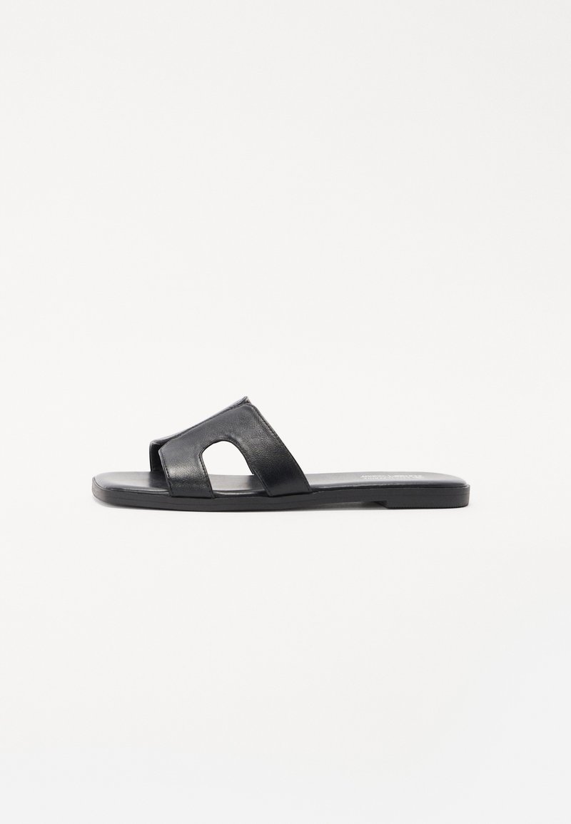 Sandale en cuir noir à enfiler avec un design découpé, semelle plate et texture lisse. Forme simple sans accents décoratifs.