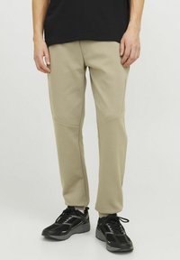 Beige sweatpants with a smooth texture, featuring patch detailing on the thighs and side pockets, paired with black athletic shoes.