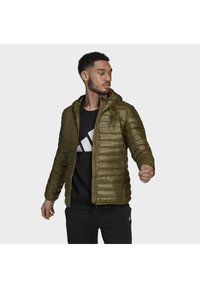 Doudoune vert olive avec un devant zippé, une capuche et un motif cousu ; portée sur un sweat noir avec un graphique blanc.