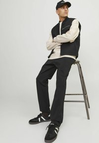 Sort og beige lagdelt outfit med en lynlåsvest, en sweatshirt, sorte bukser og sorte sneakers med hvide striber. Mand siddende på en skammel.