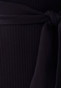 Svart ribbad tyg med en knutdetalj i midjan. Texturen är strukturerad och elastisk, med vertikala linjer och en slät yta.