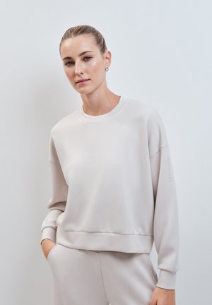 Jonge vrouw met haar in een knot draagt een lichtbeige oversized sweatshirt en bijpassende broek, staand met één hand in de zak tegen een witte muur.