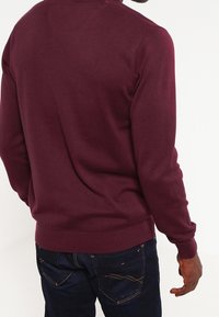 Homme portant un pull à manches longues couleur bordeaux et un jean bleu foncé, vu de dos sur un fond blanc uni.