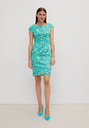 Kleid mit Kapuze in einem lebhaften Blau, verziert mit grünen und weißen abstrakten Blumenmustern. Glatte Textur, taillierte Passform, kombiniert mit leuchtend blauen Schuhen.