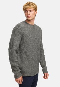 Grijze gebreide trui met een gestructureerd visgraatpatroon, ronde halslijn en geribbelde manchetten. gedragen over marineblauwe broek.