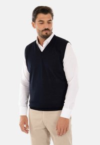 Il gilet in cotone di un blu navy presenta un design a scollo a V, indossato sopra una camicia bianca. Il tessuto è liscio e con una consistenza leggera.