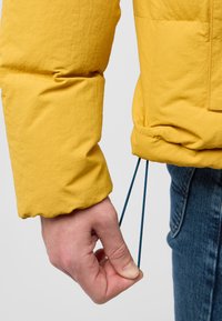 Geel pufferjack met een gestructureerde afwerking, voorzien van een trekkoord bij de pols. Een hand houdt de blauwe lijn vast, wat de pasvorm en functionaliteit verbetert.