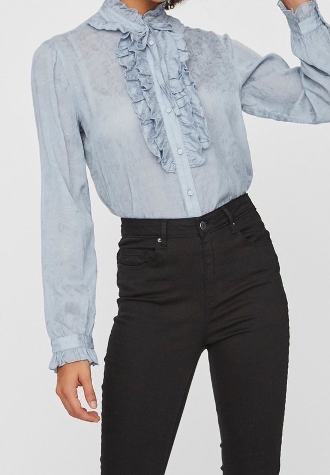 Blouse bleu clair à manches longues avec volants à l'avant et aux poignets, associée à un jean skinny noir taille haute.