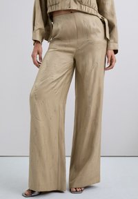Pantalones de pierna ancha en un suave tejido beige, adornados con patrones de ondas plateadas y piedras brillantes para un aspecto texturizado.