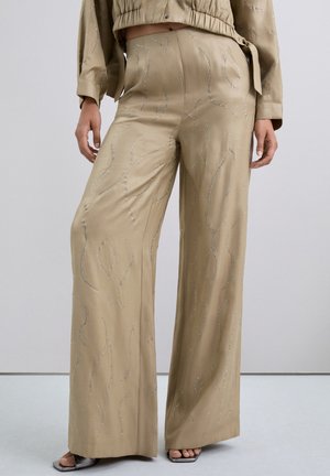 Pantalones de pierna ancha en un suave tejido beige, adornados con patrones de ondas plateadas y piedras brillantes para un aspecto texturizado.