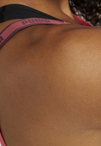 Czarny regulowany pasek z różowo-bordowym logo PUMA, ukazujący gładki brązowy odcień skóry na ramieniu oraz subtelną teksturę.