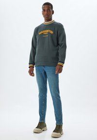 Sudadera verde oscuro con el texto amarillo "WASHINGTON STATE", cuello de canalé con franjas amarillas y blancas. Jeans azul claro y botas verdes.