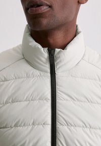 Jasnoszara kurtka puchowa z wysokim kołnierzem, poziomym pikowaniem i kontrastującym czarnym zamkiem błyskawicznym. Gładkie wykończenie tkaniny.
