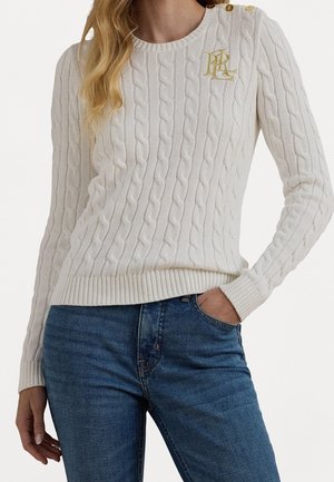 Femme portant un pull en maille torsadée blanc avec des boutons dorés et un logo sur l'épaule, associé à un jean en denim bleu, main dans la poche.
