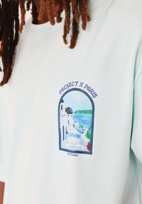 Camiseta de manga corta azul claro con un estampado gráfico de un paisaje de Santorini que incluye arcos, cúpulas azules y vegetación.