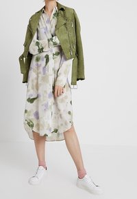 Grön mockajacka i cropped stil över en skjortklänning med blommönster i pastellfärger, parat med vita sneakers och ljusrosa strumpor.