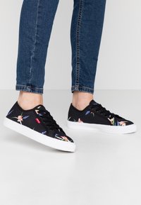 Person klädd i blå jeans och svarta sneakers med vita sulor och färgglada blommönster som står på ett vitt golv.