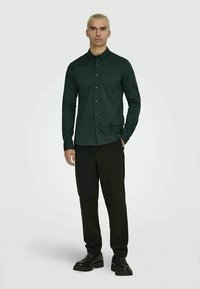 Homem com cabelo loiro curto, vestindo uma camisa verde-escura de botão, calças pretas e sapatos pretos, em pé contra um fundo branco liso.