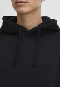 Sudadera negra de algodón con una textura suave, que cuenta con un bolsillo tipo canguro en la parte delantera, cordones y un sutil logo en relieve en el pecho.