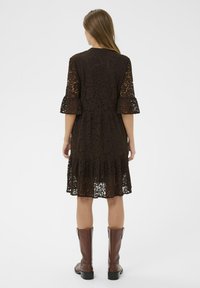 Ruda nėriniuota suknelė su gėlių raštais, platėjančiomis rankovėmis ir gėlėmis puošta apatine dalimi. Sujungta su kelių aukščio rudais batais, sukuria sluoksniuotą įvaizdį.