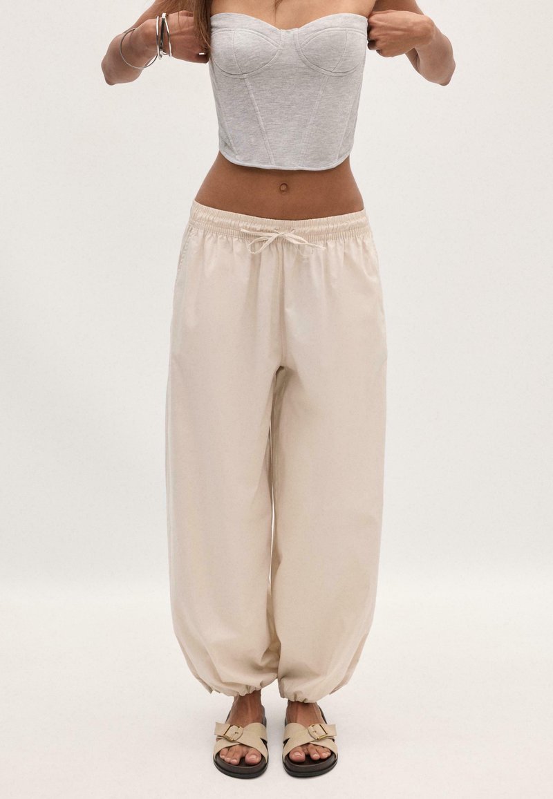 Femme portant un pantalon jogger beige avec cordon et poignets élastiques, un haut corset gris sans bretelles, des bracelets argentés et des sandales beige avec boucles.