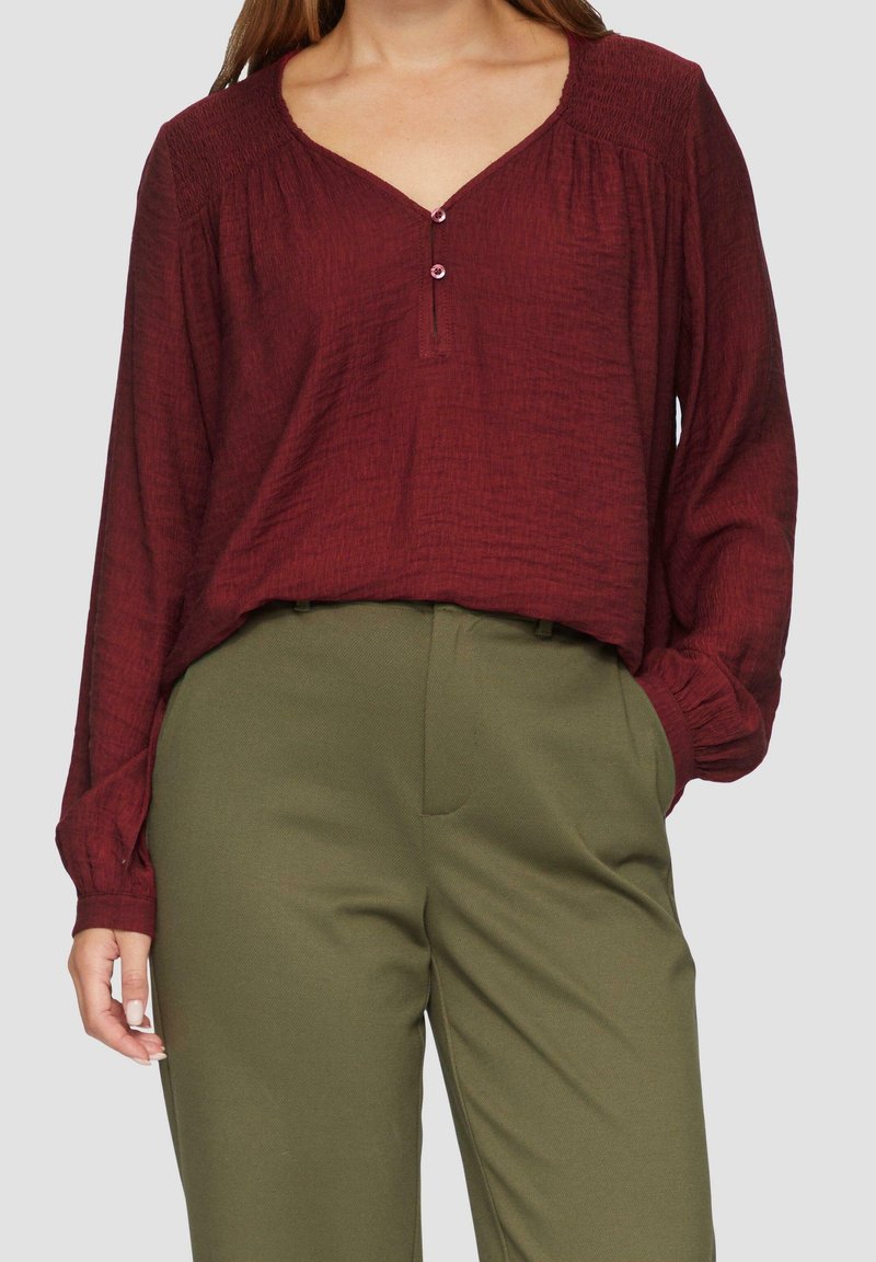 Femme portant un chemisier bordeaux à manches longues avec deux boutons et un pantalon vert taille haute, main dans la poche sur un fond uni.