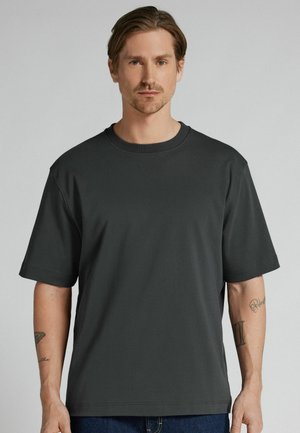 Mężczyzna o średniej długości włosach, ubrany w luźną ciemnoszarą koszulkę i niebieskie dżinsy, stojący na tle jednolitego jasnego tła.