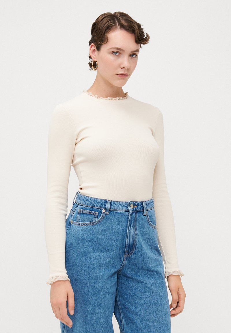 Haut à manches longues crème avec un col volanté, associé à un jean bleu taille haute. Texture lisse, design ajusté, style décontracté.