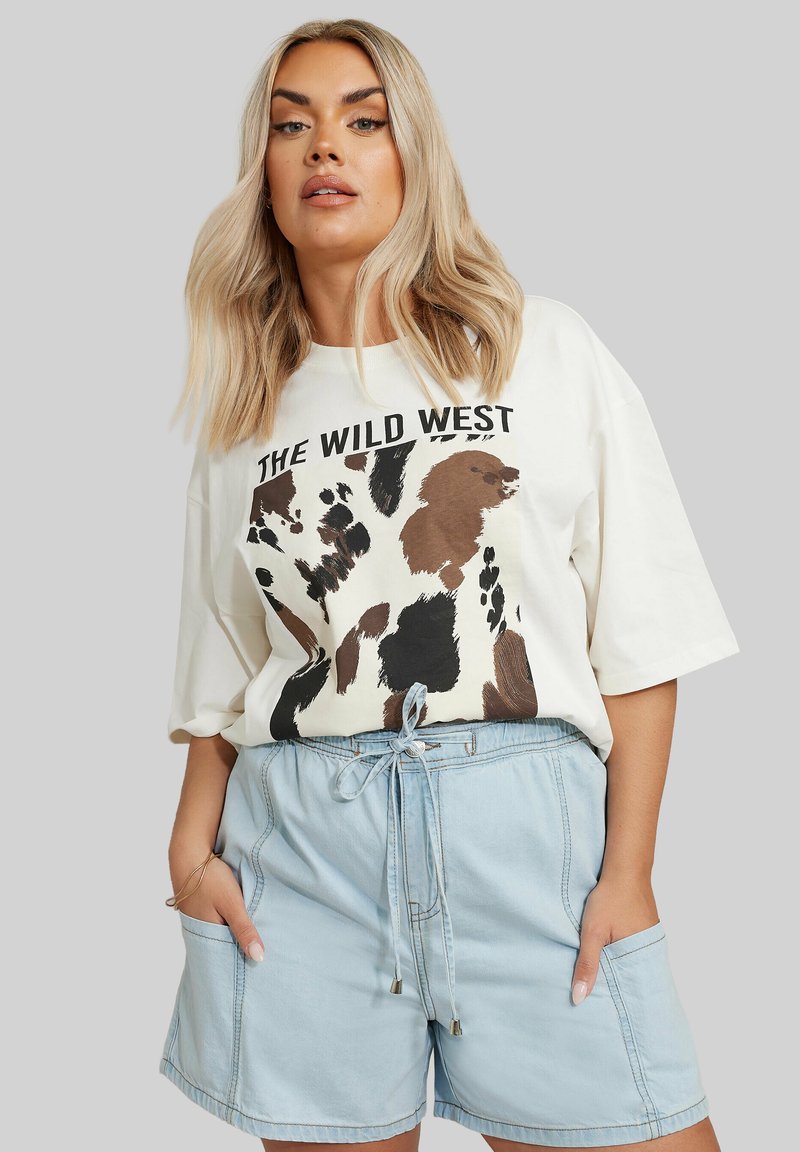 Valkoinen ylisuuri t-paita, jossa on teksti "THE WILD WEST" ja ruskea eläinkuos, yhdistettynä vaaleansinisiin nippanappa-housuihin, joissa on etutaskut.