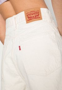 Shorts en denim blanc taille haute avec un patch en cuir Levi Strauss & Co., présentant une couture de poche arrière dans un motif d'arc distinctif.