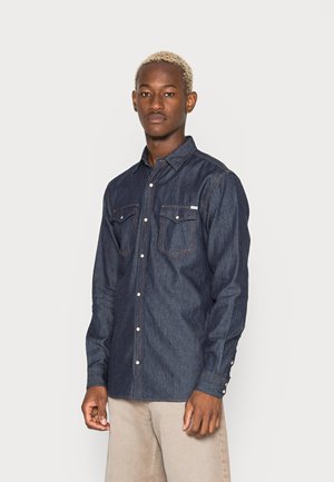 Camisa de botón de mezclilla en azul oscuro con dos bolsillos en el pecho, costuras naranjas en contraste y botones perlados. Cuello clásico y mangas largas.