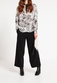 Blusa macchiata in bianco e nero con colletto a fiocco, abbinata a pantaloni neri a gamba larga e stivaletti neri, con una pochette beige.