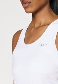 Cămașă sport albă cu decolteu rotund, textură netedă, având un etichetă cu brandul "TED BAKER SPORT" în negru și roșu.